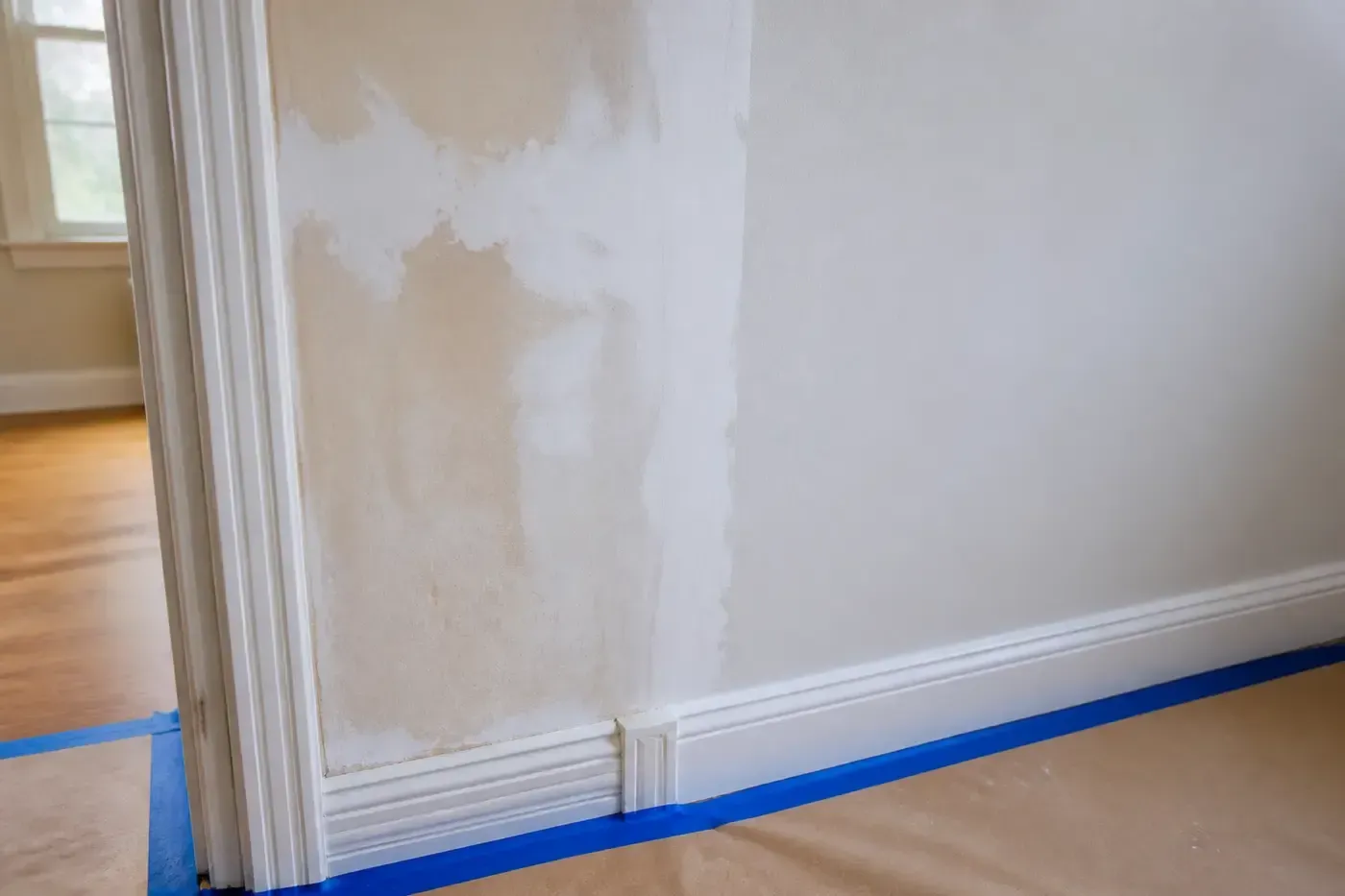 A prepared interior wall shows repaired plaster and a smooth primed surface beside protected trim and flooring.